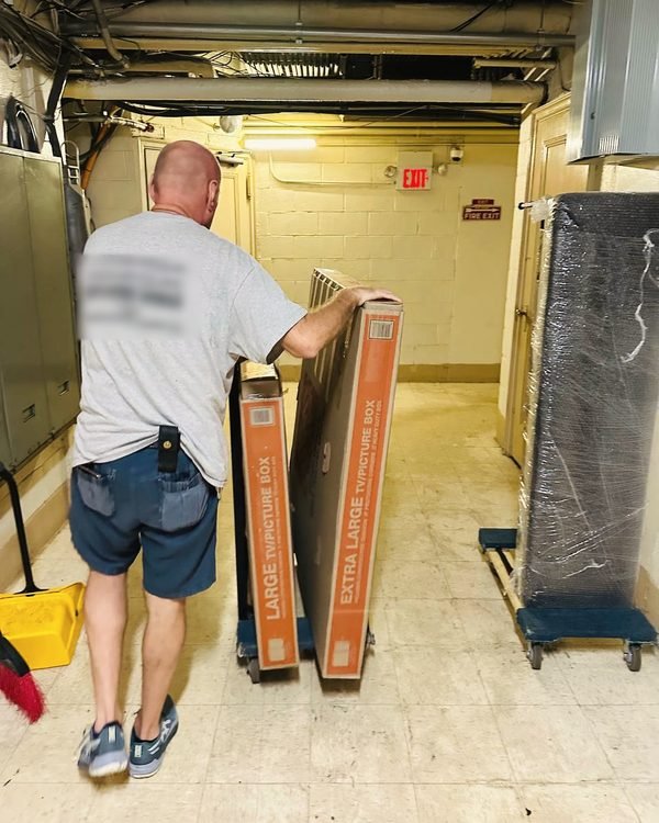 A mover wheeling large TV and picture boxes through a building basement corridor
