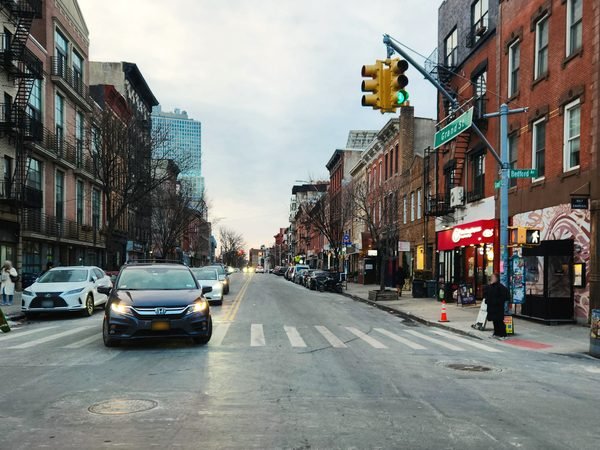 A busy corner in Williamsburg, Brooklyn: walk-up buildings with fire escapes, a crosswalk, traffic